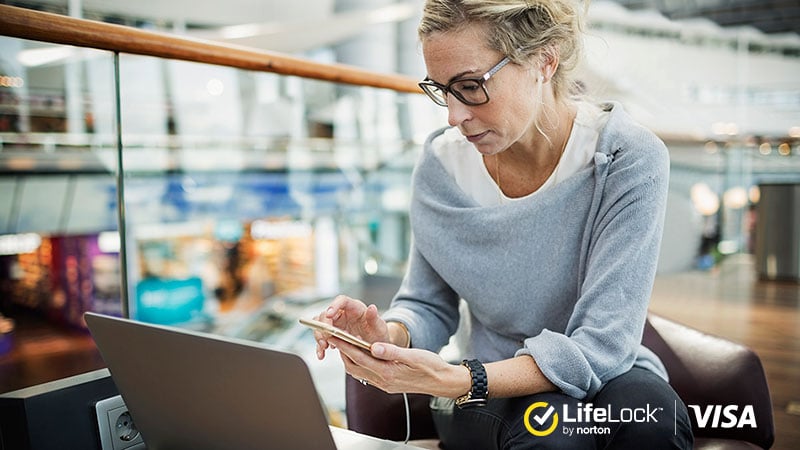 Woman using phone and laptop with LifeLock and Visa logos.