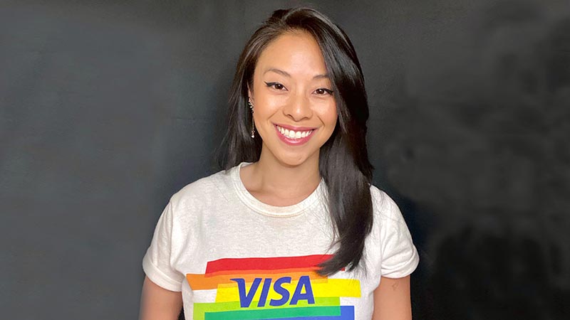 Woman wearing a Visa tshirt with the pride flag, smiling at the camera in front of a gray background.