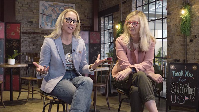 two women (Amy and Vivi from pig and a jelly jar) sitting in cafe, addressing camera