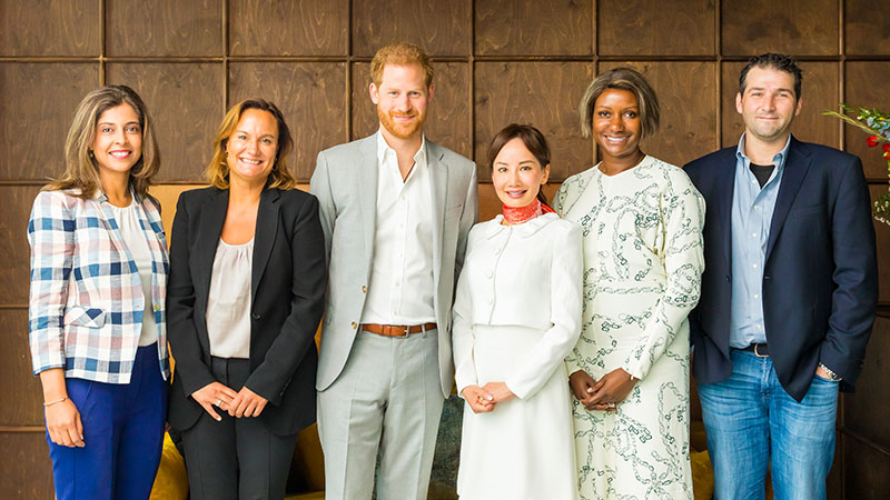 Group of panel speakers, including HRH The Duke of Sussex, smiling and standing against a brown wall