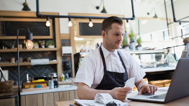 Man in an apron working on a laptop at the front counter of his shop. Man in an apron working on a laptop at the front counter of his shop.
