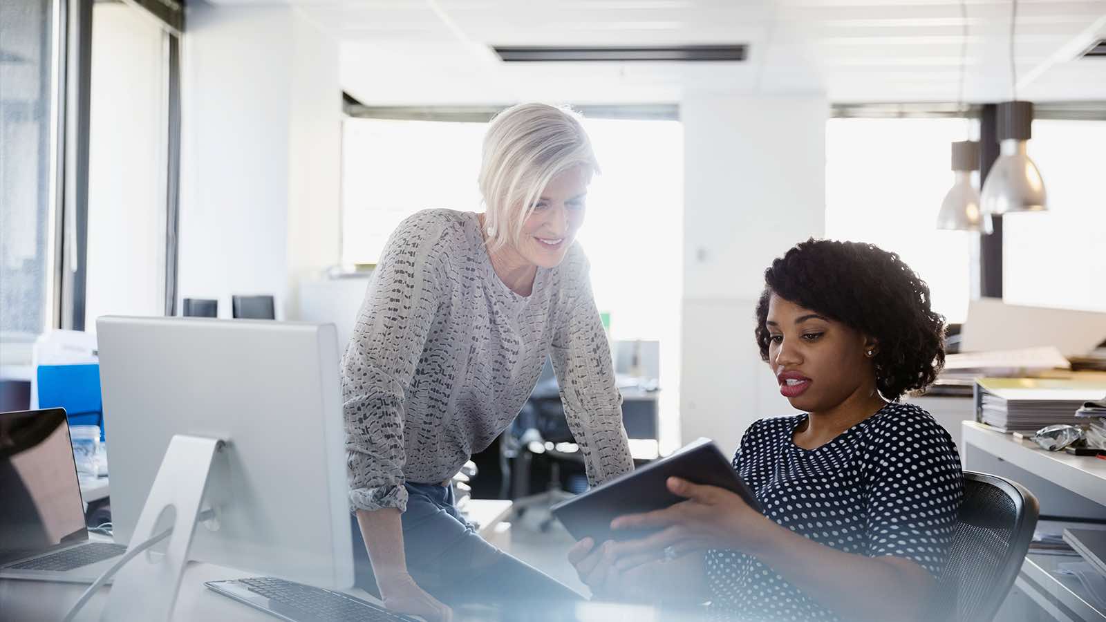 A woman sitting at a desk while holding a tablet and showing it to another woman.