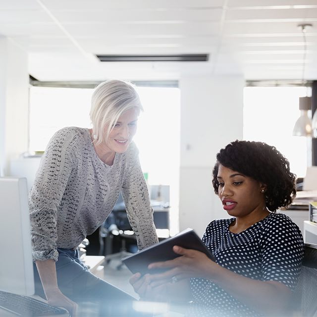 A woman sitting at a desk while holding a tablet and showing it to another woman.