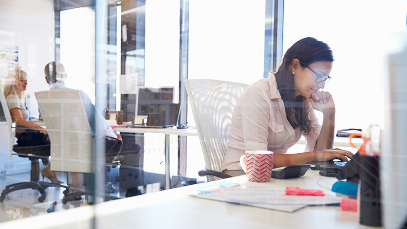 Woman working at her desk in an office with people in the background chatting at their desks. Woman working at her desk in an office with people in the background chatting at their desks.