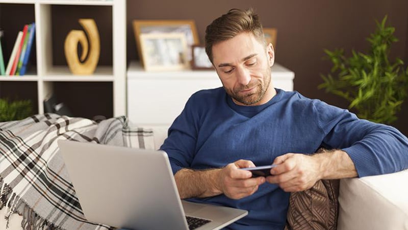 Man sitting on a couch with a laptop on his lap holding a smart phone with both hands.
