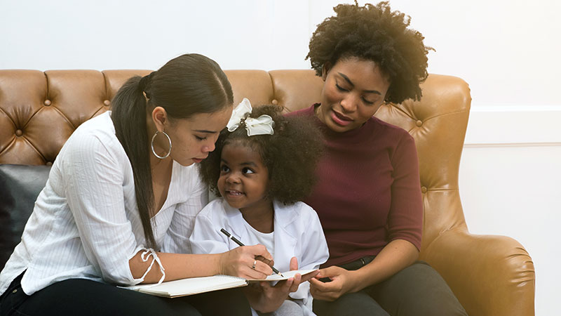 A Black female LGBT couple and their child sit on the couch working on her homework.