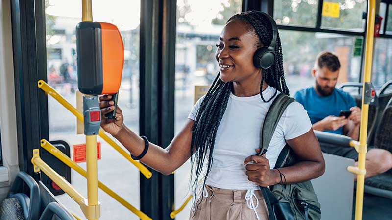 A woman, smiling and wearing headphones, taps her card to pay her fair on a city bus