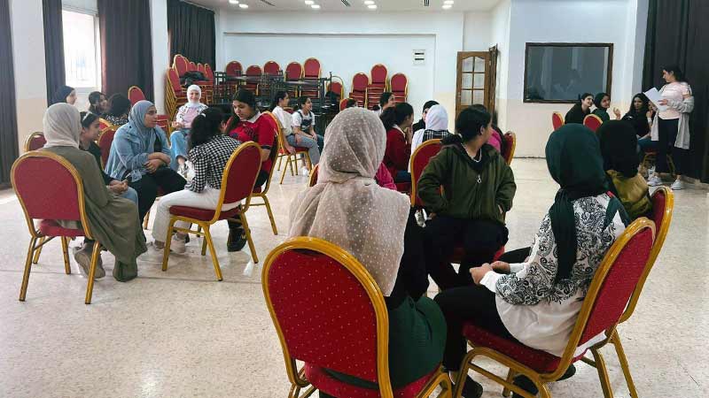 Girls and women sit around tables in a room listening to a speaker.