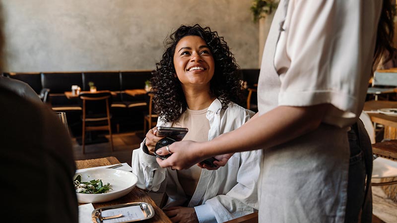 Woman using smartphone while paying restaurant bill with contactless payment during dinner party with friends