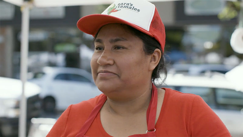 Small business owner Sara Rodriguez smiles off camera wearing a hat with her company's logo.