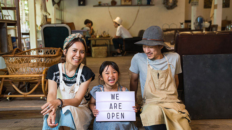 A couple sits outside their shop with their young daughter between them smiling and holding up a sign indicating the store is open. A couple sits outside their shop with their young daughter between them smiling and holding up a sign indicating the store is open.