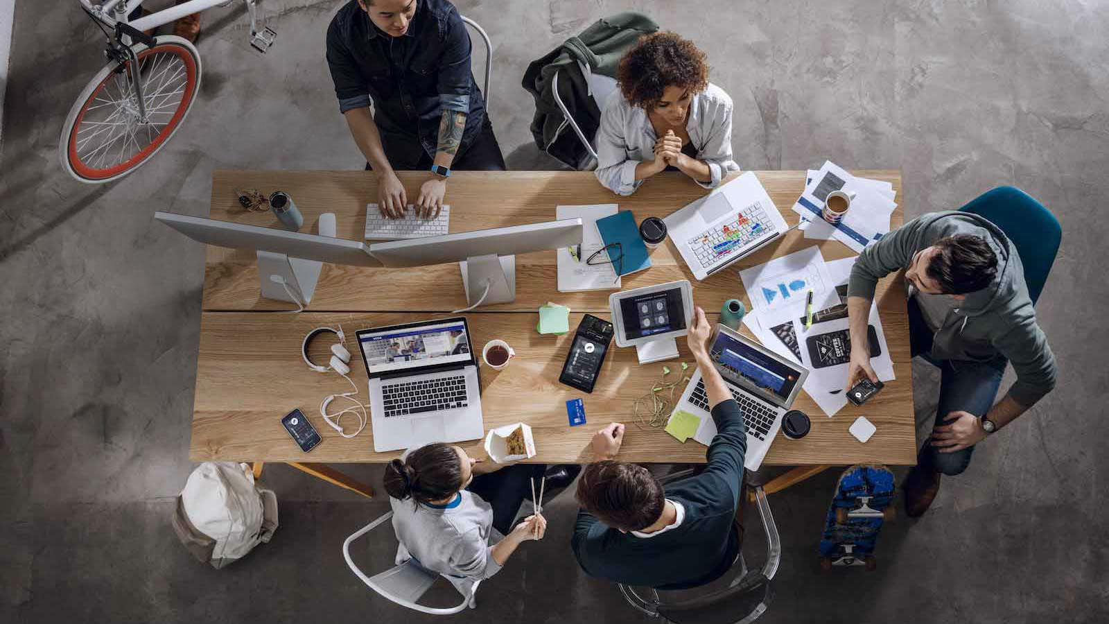 Overhead view of employees at a table with laptops, coffee mugs, notebooks, and paper spread out.