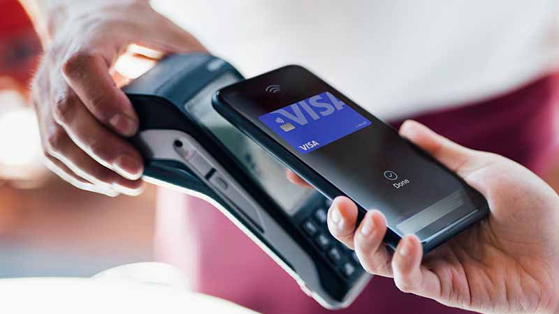 Hand holding a phone showing the image of a Visa-branded card up to a payment terminal.