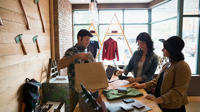 Customer using credit card reader at clothing shop counter, while man behind shop counter holds bag of her purchases. Customer using credit card reader at clothing shop counter, while man behind shop counter holds bag of her purchases.
