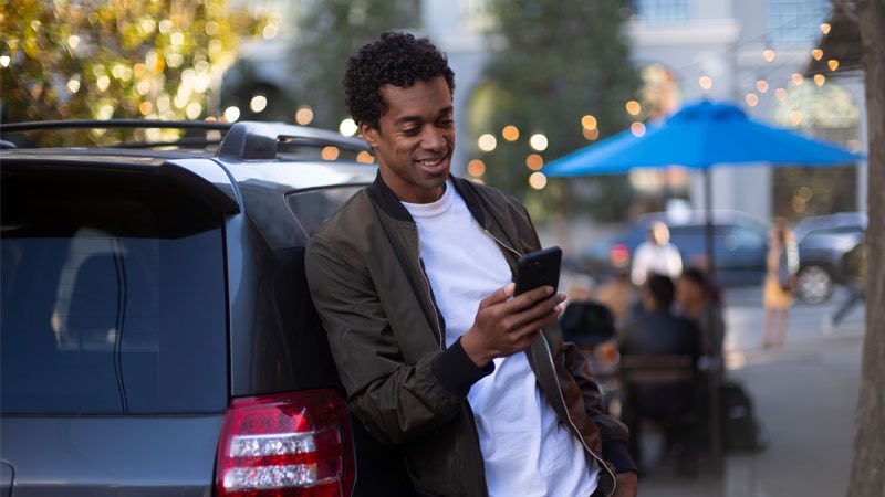 Smiling man leaning against car, looking at his phone.