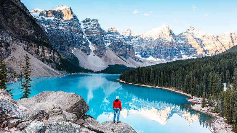 Man standing on rock, looking at a lake and snow-capped mountains.